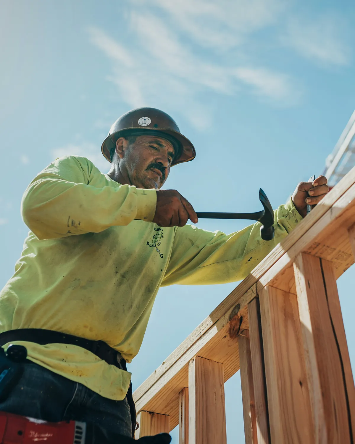 A framing carpenter in a hard hat and work shirt, hammering at a timber stud wall