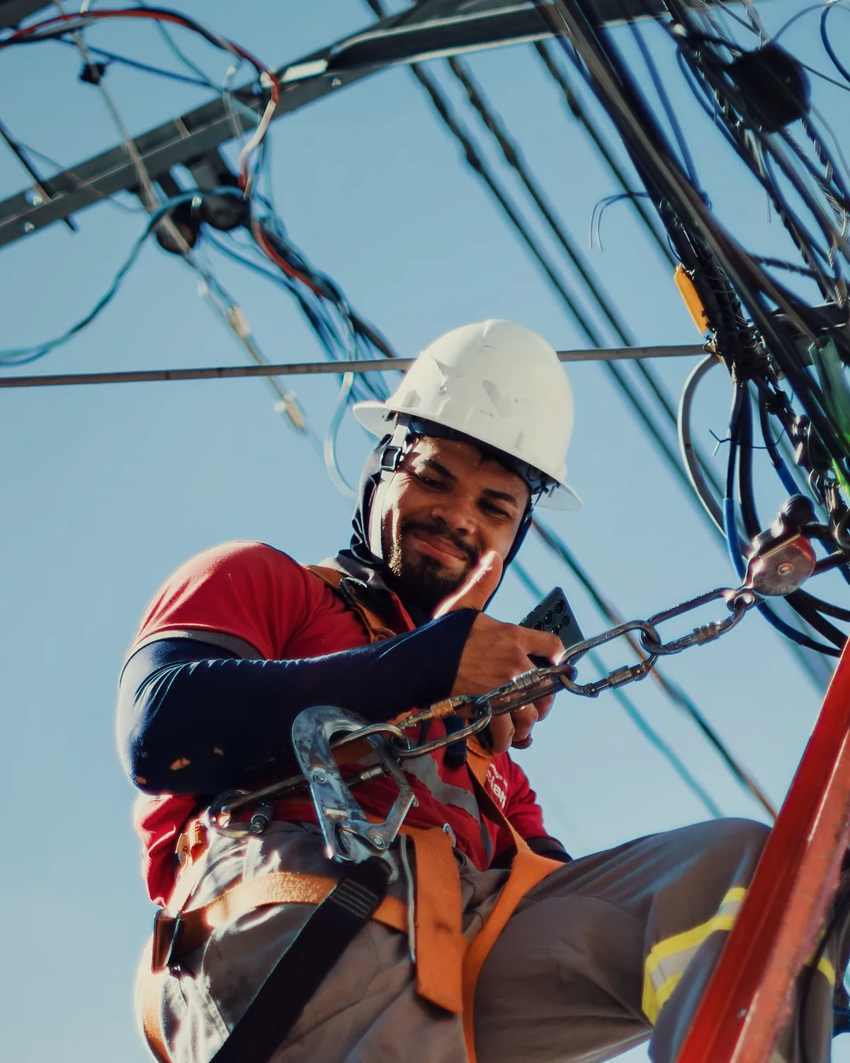 A lineworker in a hard hat and harness, smiling, high on a pole among the lines